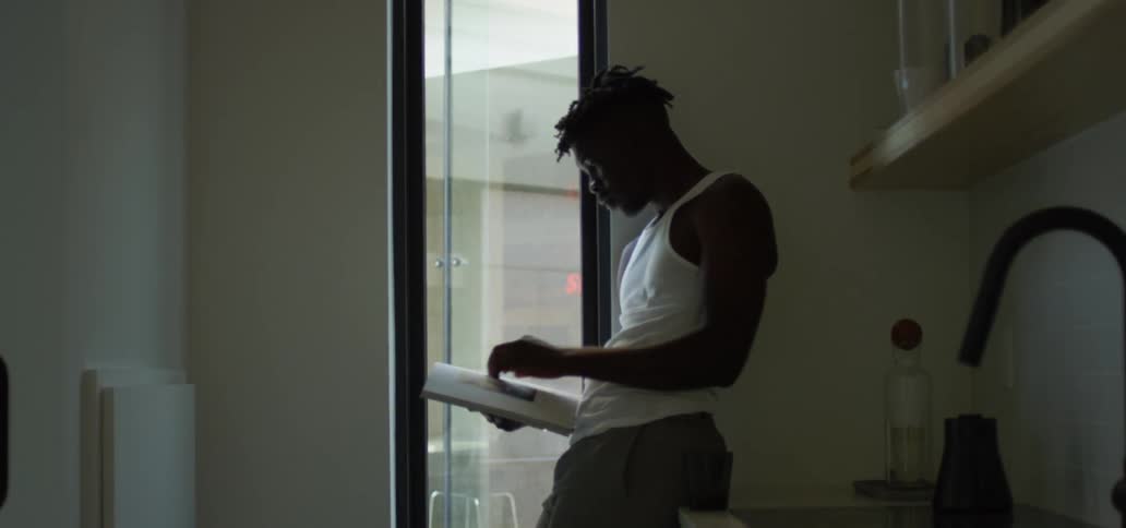 Male leans against the kitchen counter next to a window while taking a drink from a cup before placing the cup on the counter and turning the page on the book he is reading