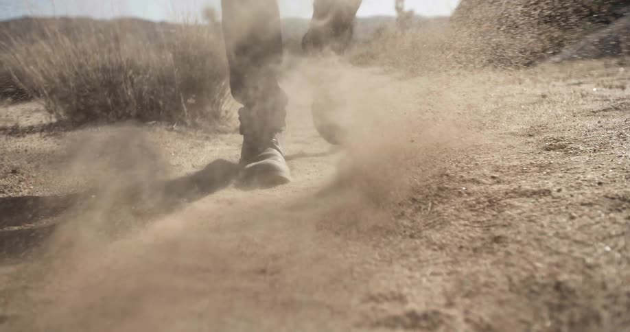A man is walking in a dry canyon and kicks dirt toward the camera