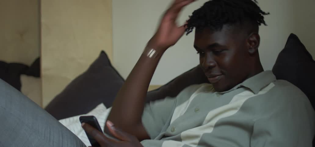 Black man with a grey polo shirt and pants sitting on bed smiles and uses a smartphone in the bedroom