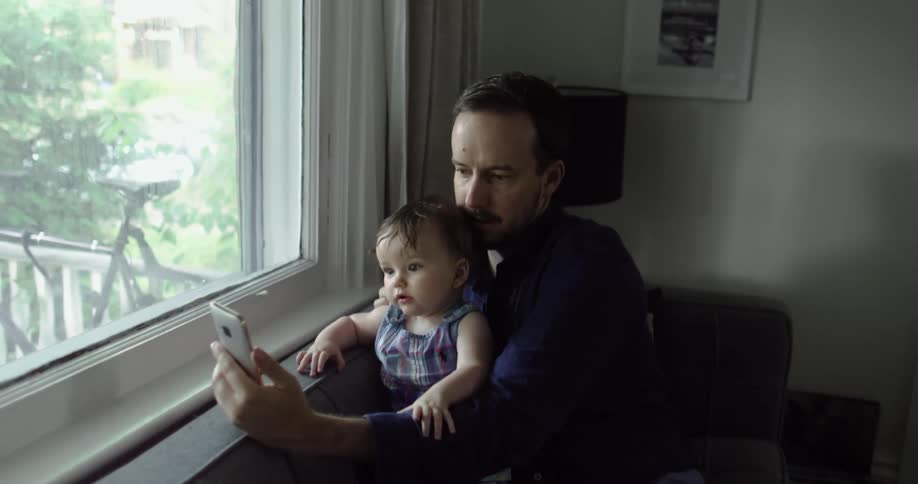 Father sitting on couch near a large window extends his smart phone out in front of baby daughter while video chatting