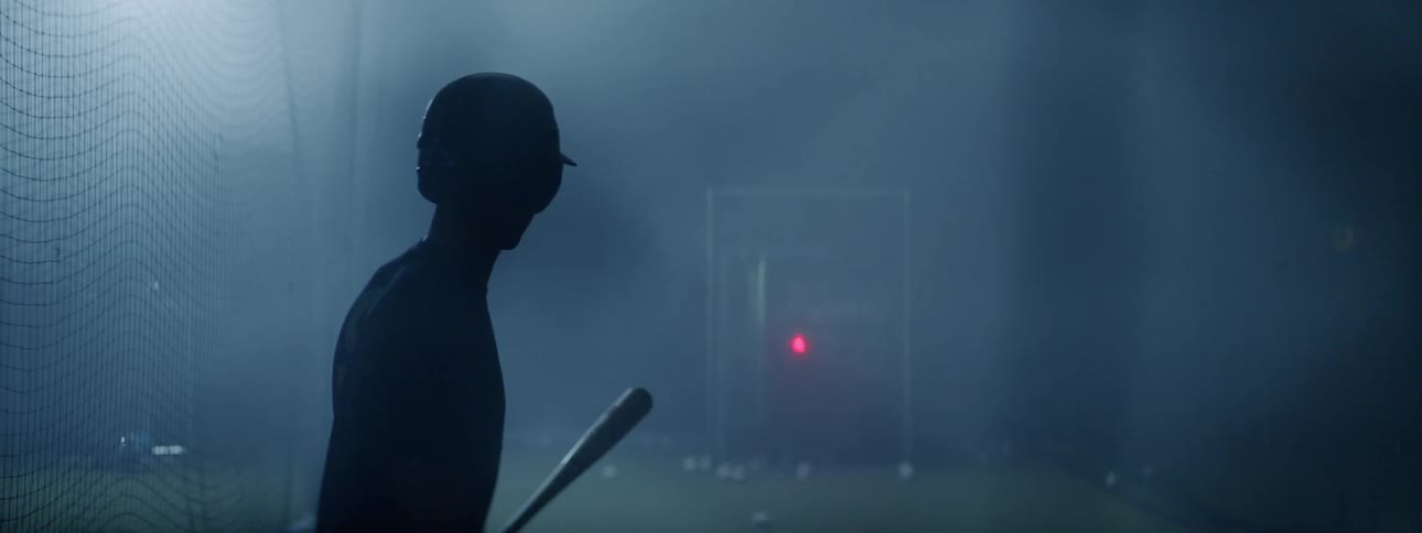 Young man enters a batting cage holding a baseball bat then a pitching machine pitches a ball towards him