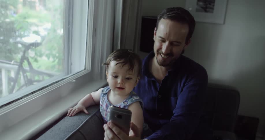 A father takes a selfie with his infant son as they sit on a chair next to a large window