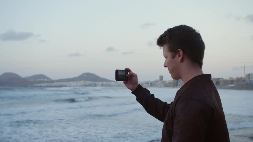 Young man films ocean with smartphone at sunset