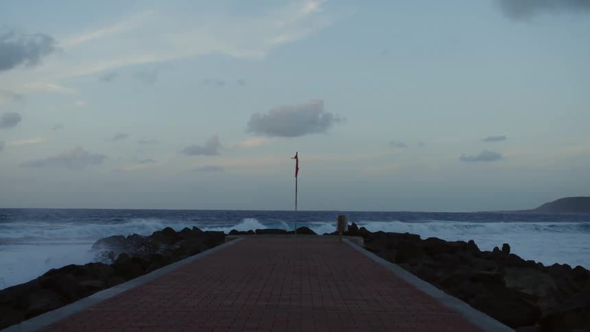 A flag flaps in the wind on a brick paved walkway that is surrounded by rocks with crashing waves in the background at dusk