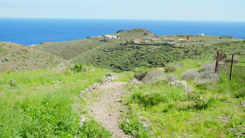 Dirt path leads past fence toward compound occupying hill near ocean