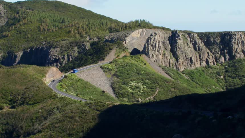 A blue bus and two cars drive on a mountain highway in the distance