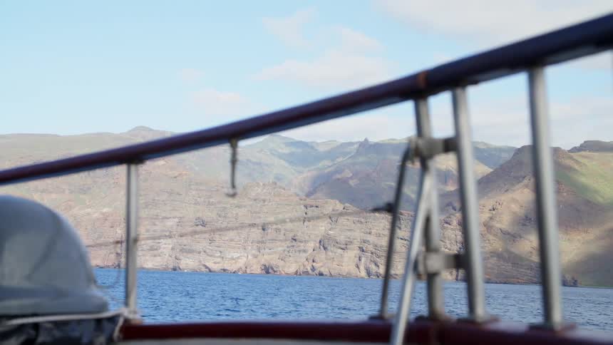 Boat bobs along ocean paralleling cliff on shore backed by mountains
