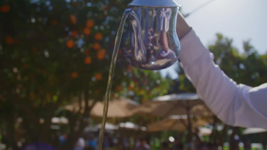 A waiter holds a silver kettle up high as he pours a drink on a restaurant patio