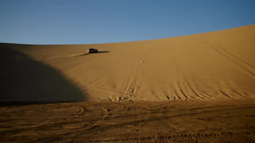A sport utility vehicle drives up the side of a tall sand dune