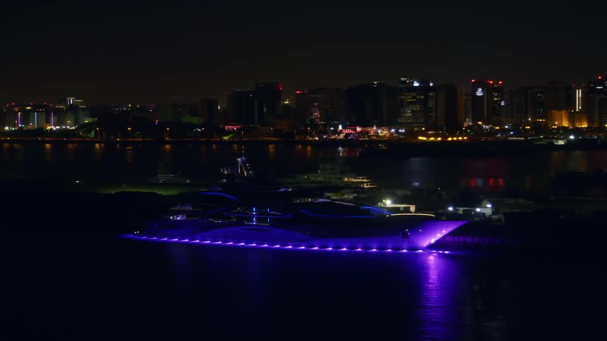 A yacht is lit underneath with bright purple lights as it travels slowly through the ocean in front of a city at night