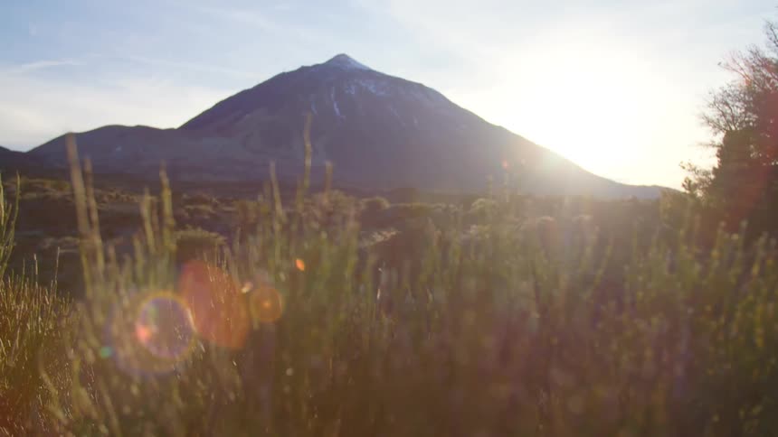 Sunlight peaking from behind a distant mountain shows onto the swaying grass of a countryside