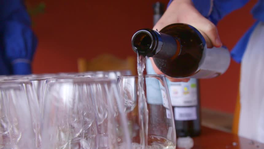 A waitress pours white wine into several wine glasses lined up on a table