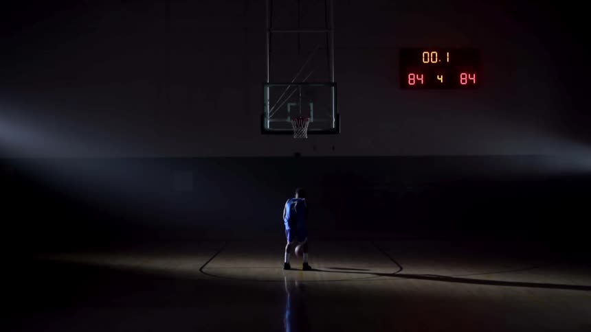 A teenager in a dark gym dribbles a basketball and then shoots it in to the basket
