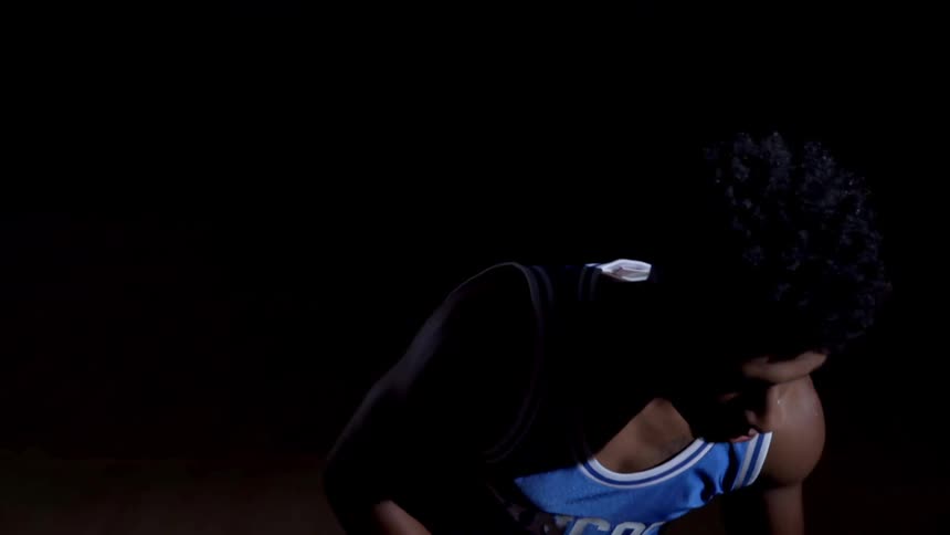 A basketball player wearing his school jersey stands on a dark court dribbling a ball