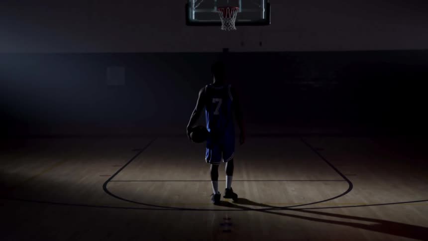 A teenager walks on to a dark basketball court and dribbles a ball