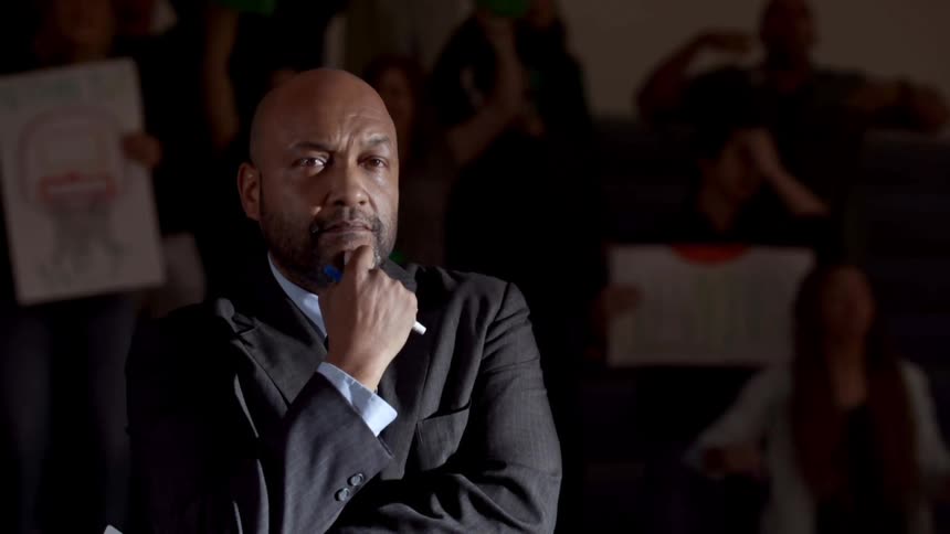 A black man in a suit sits with a stern look on his face in front of a group of yelling teenagers holding signs