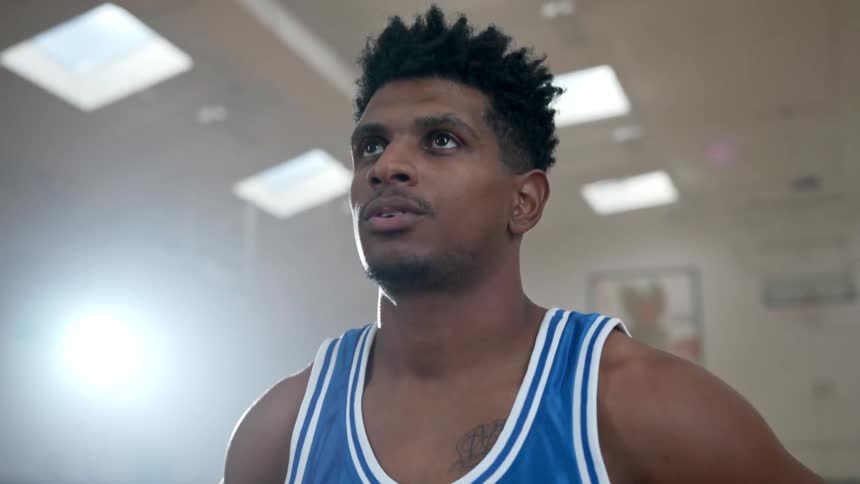 Black basketball player with a blue jersey stares up at a basketball hoop in a basketball court