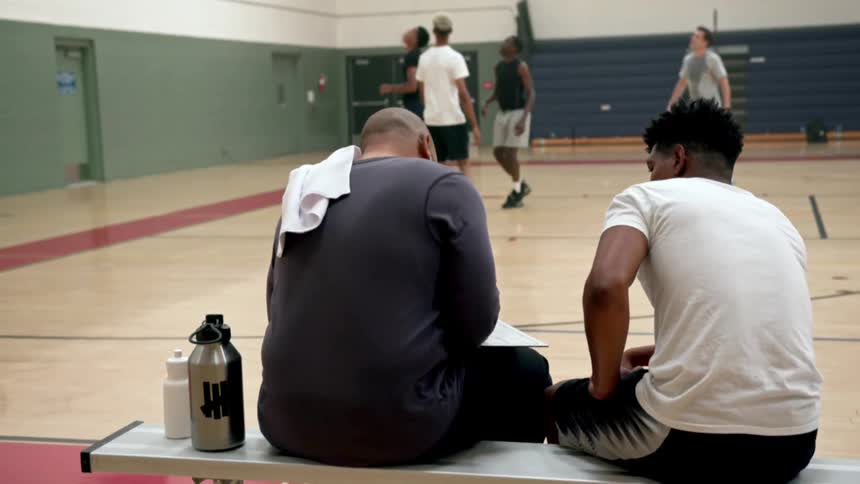 Two men sit on a bench in the gym while four other men practice playing basketball