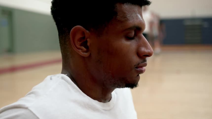 Sweaty black basketball player with a white t shirt sits on a bench eyes closed and breathes out in a basketball court