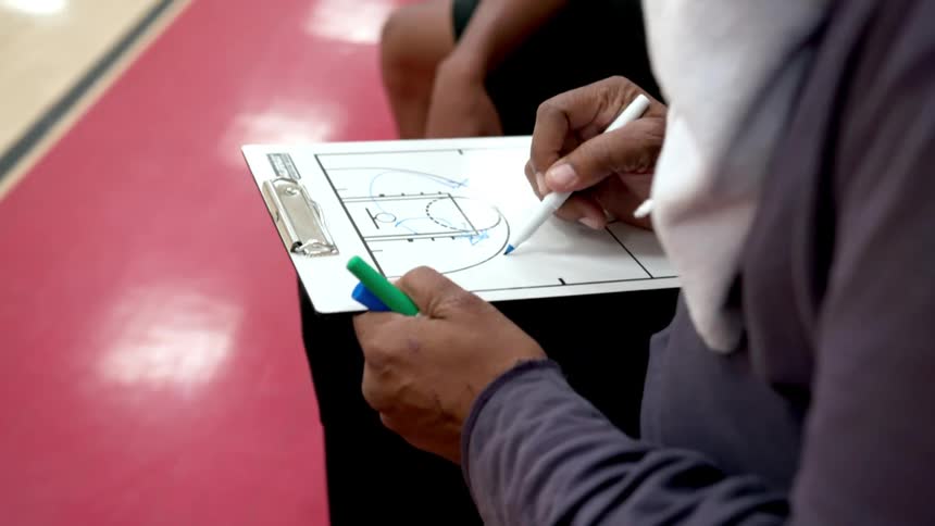 A coach diagrams a play on a board for an athlete sitting next to him on the bench of a basketball court