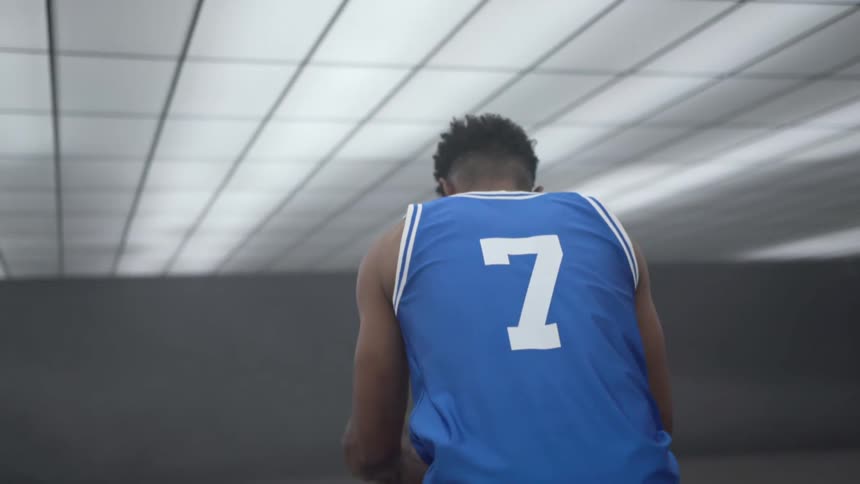 As the lights come on in a gym a teenager dribbles and prepares to shoot a basketball