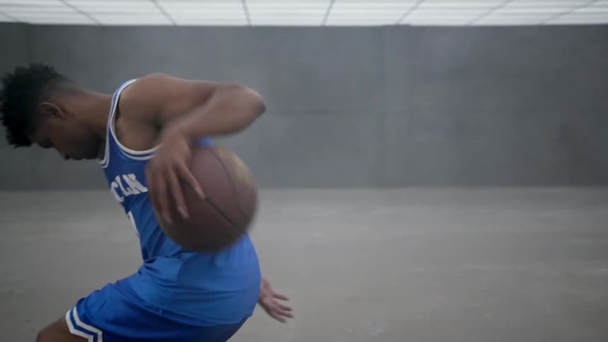 A black male teenager in a basketball outfit dribbles a ball in a room