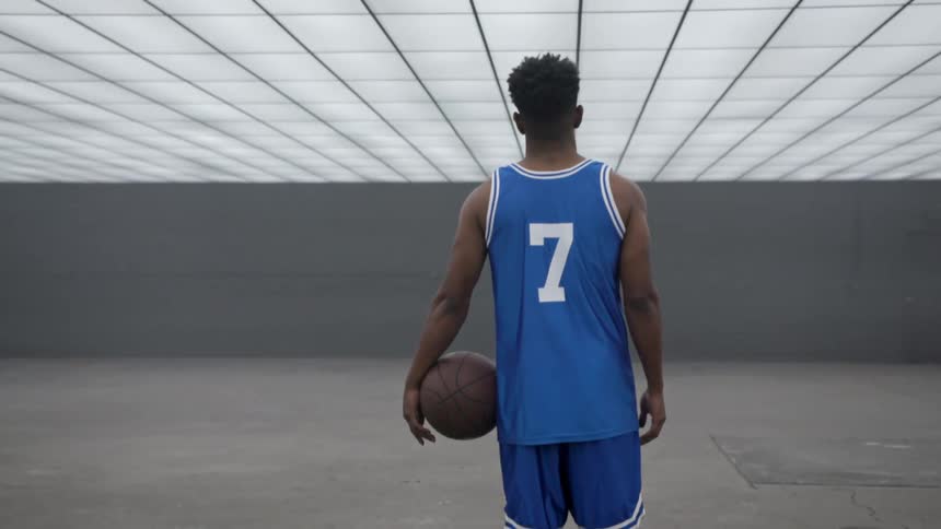A black man in a basketball uniform holds a basketball while standing indoors