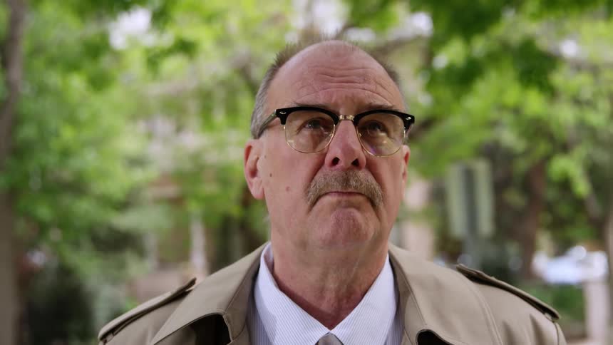 Elderly man with a trimmed mustache and a trench coat raises his head