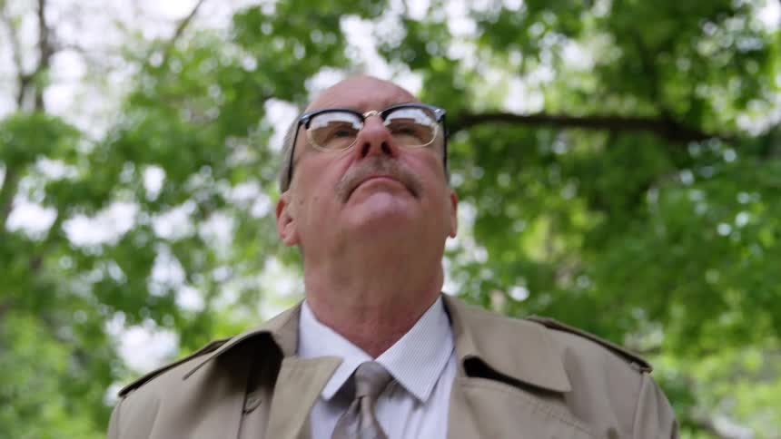 Elderly family doctor with a trimmed mustache and trench coat walks in the public park
