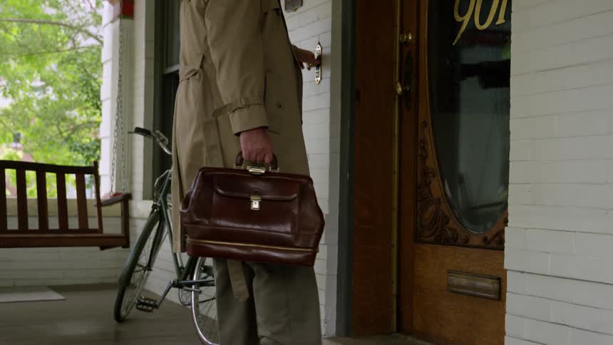 Elderly man with a trench coat and carrying a brown bag walks up to the door to ring the bell