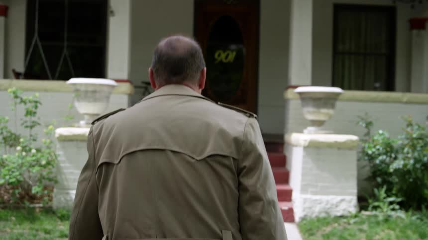 Man wearing a coat walks through a gate and up the steps towards a white house