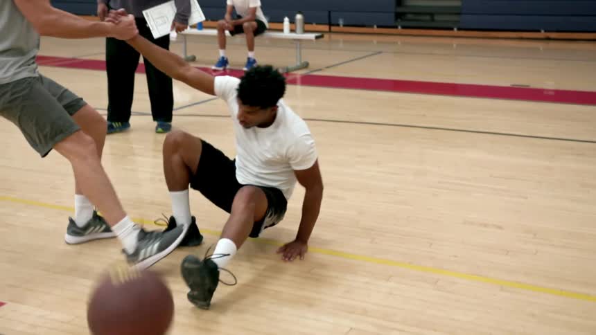 A coach watches as one team mate helps a fallen team mate up off the basketball court