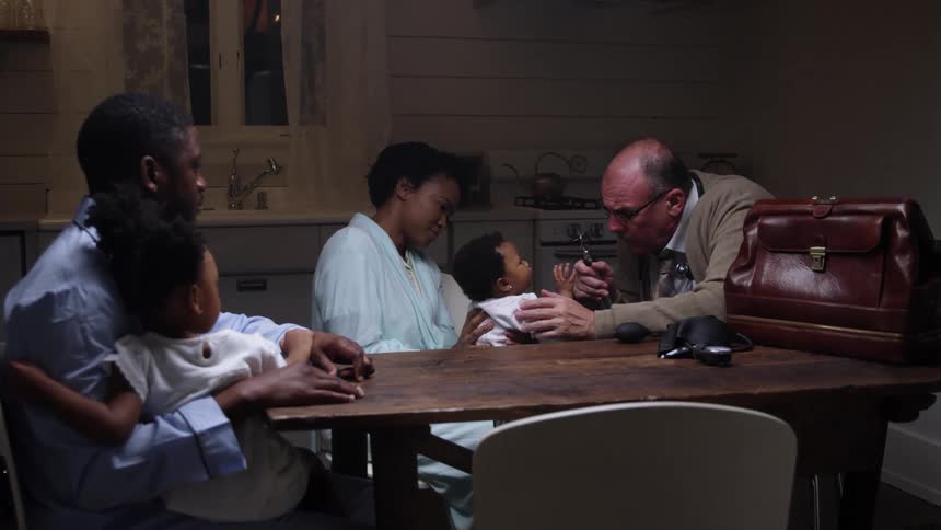 A doctor visits a family in their home and examines a baby girl as the mother holds her and the father holding another child watches