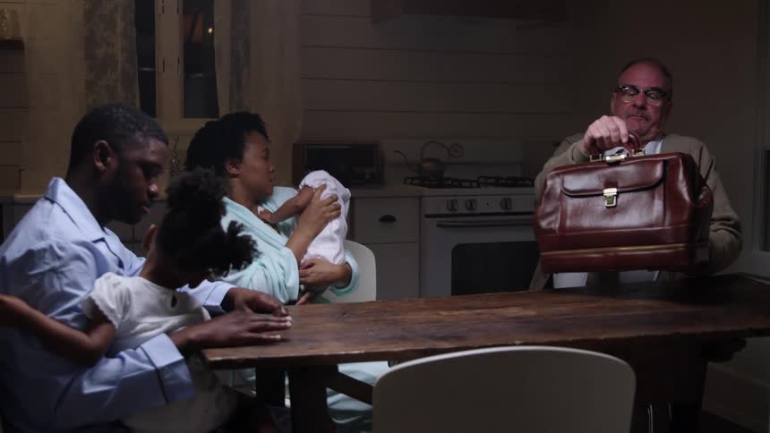 A woman sits down at her kitchen table with her baby as a doctor sits to examine her