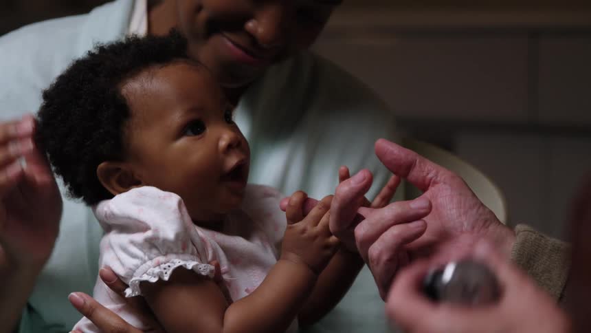 Daughter sits on mothers lap as the baby is holding on to the doctors finger while she looks up and smiles at him
