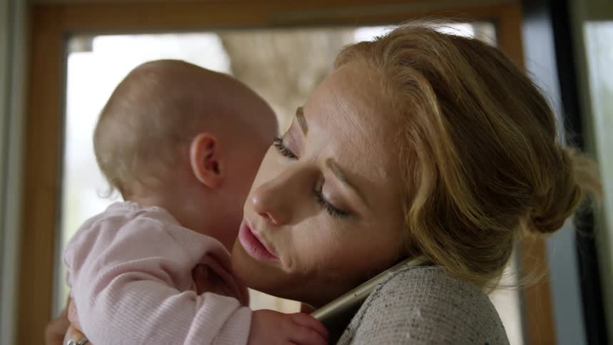 A woman holds a baby while cradling a cell phone between her ear and shoulder
