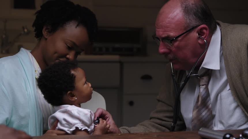 A mother holds her baby as a doctor listens to her heart during a home visit