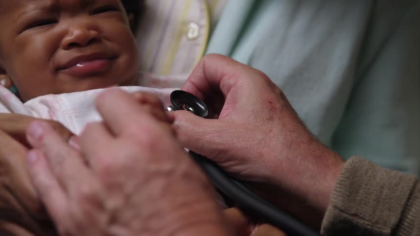 An infant girl cries and pushes away as a doctor uses a stethoscope on her chest