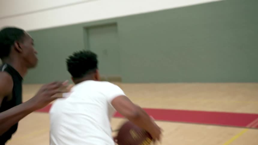 Black basketball player with a white shirt dribbling the ball bouncing it up and down on the floor drives in to shoot in a basketball court