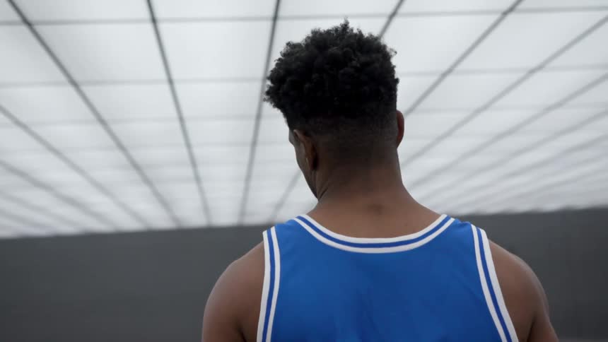 A black man in a basketball jersey stands indoors and turns his head