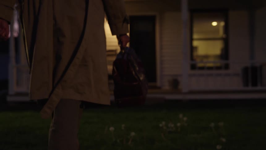 A family doctor wearing a trench coat and carrying a leather bag walks through the yard of a house toward the front door