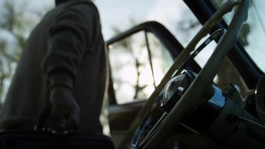 Elderly family doctor with a trench coat and holding a brown bag gets out of a classic car