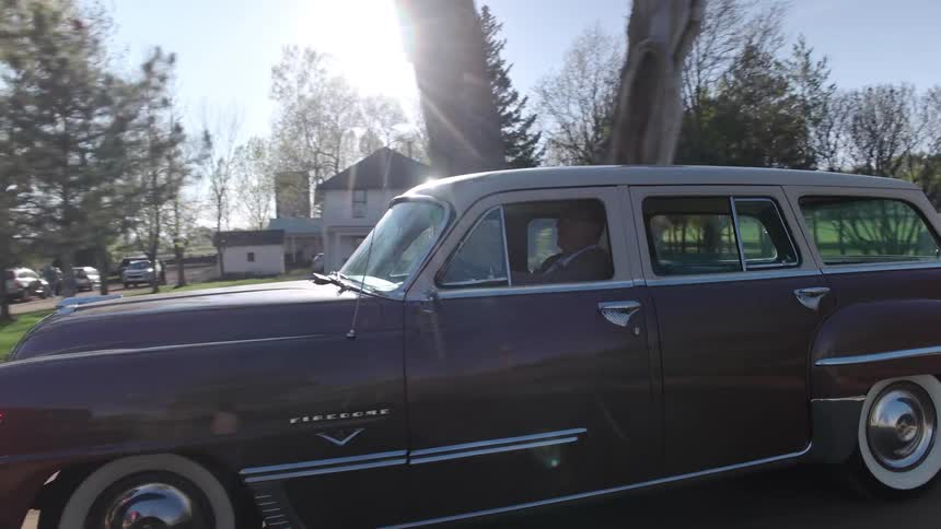 A man drives a classic car down a dirt road past houses in a rural area