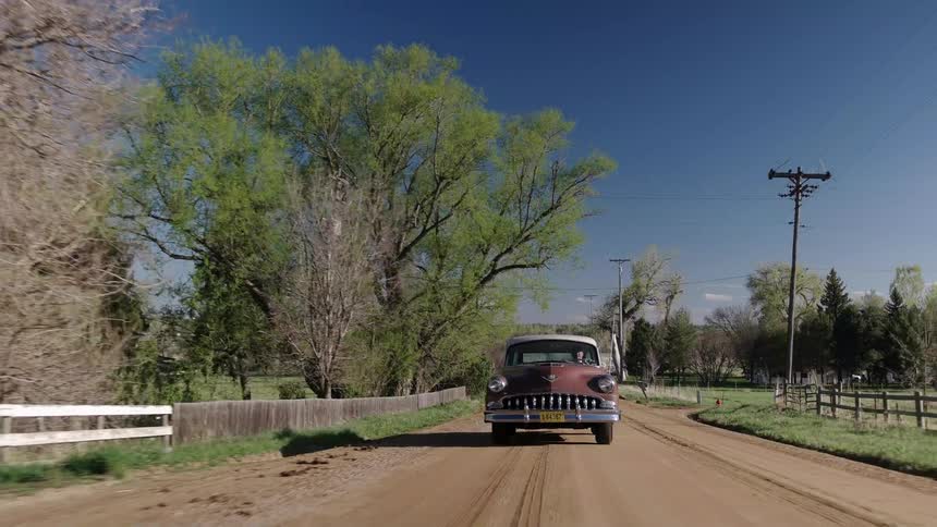 A vintage car travels along a dirt country road