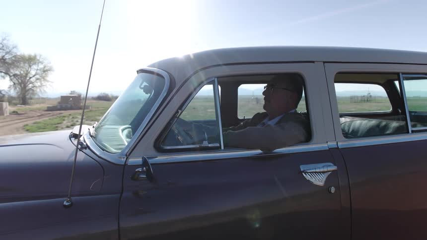 Elderly man with a trimmed mustache and a trench coat drives on highway in the countryside