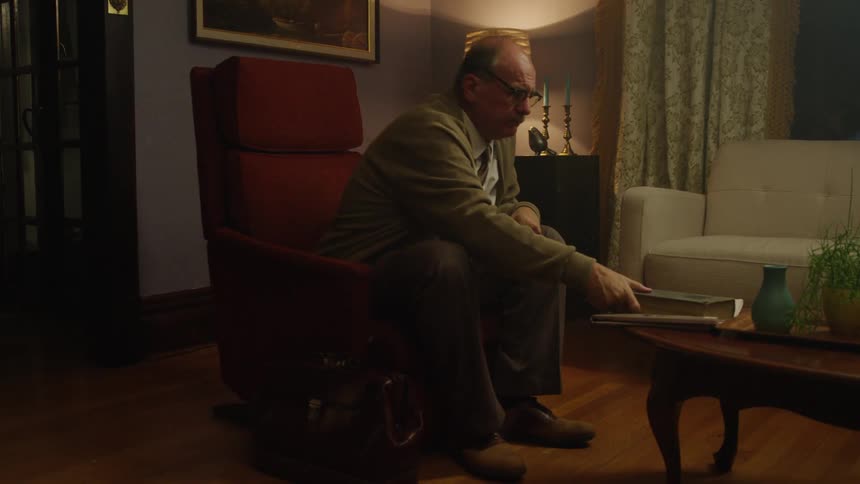 An older white man reads a book in a chair and then gets up and walks away