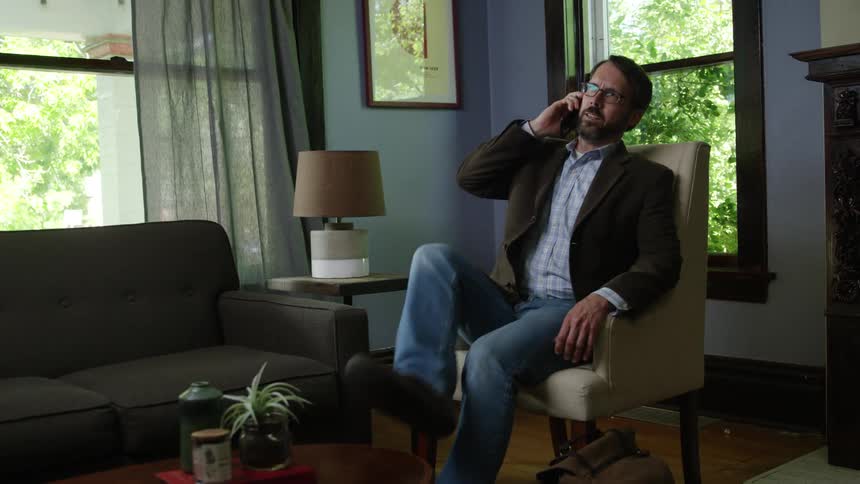 A man talking on his cellphone walks in to his living room and sits down
