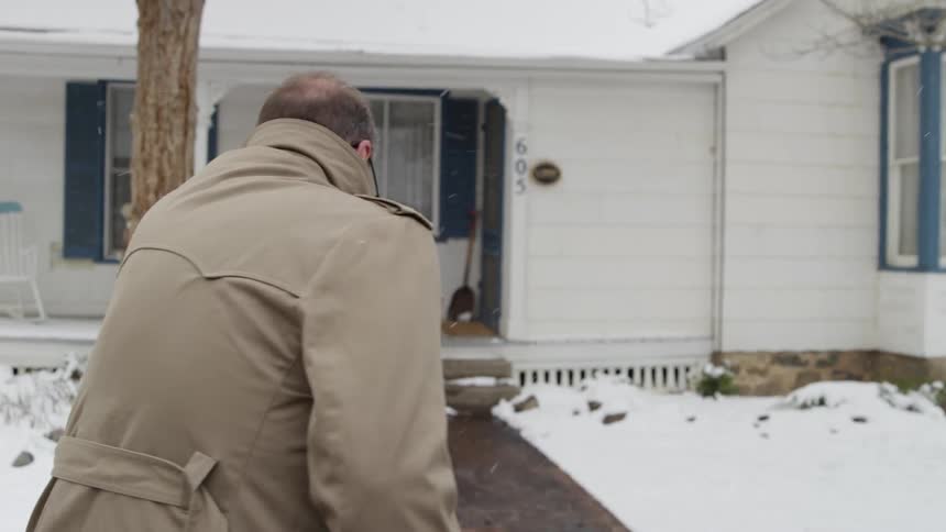 A family practitioner with glasses and a trench coat walks up to a house as it snows