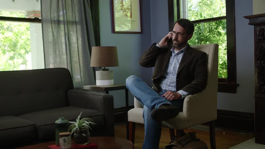 Male sitting cross legged in a chair in the living room while talking on his smart phone looks anxious after finishing his conversation