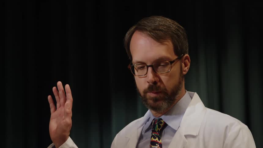 A doctor in a lab coat raises his hand while talking
